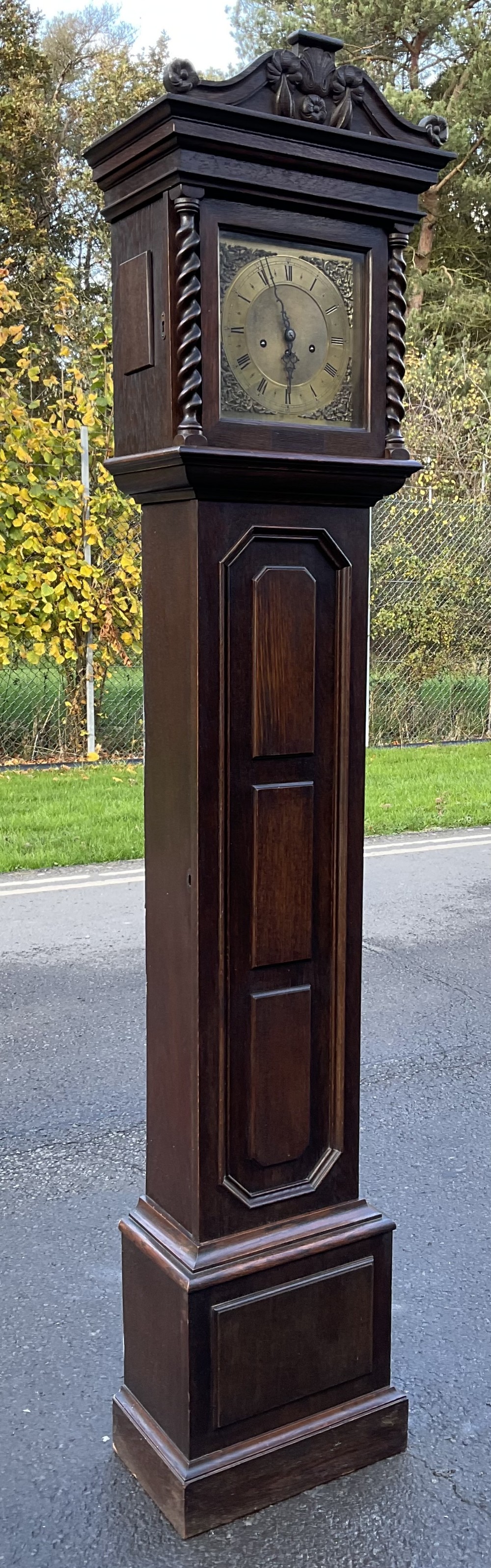 A dark oak longcase clock featuring a carved pediment, barley twist columns, a brass dial with decorative spandrels, and geometric panel moulding on the trunk door.