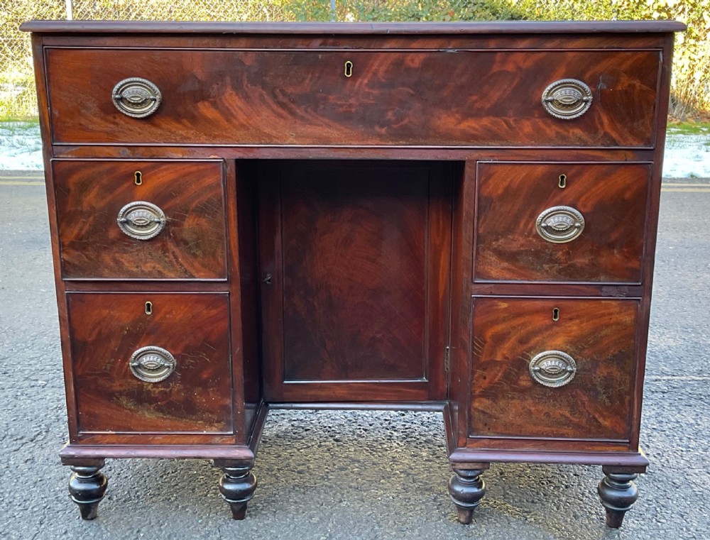 A Georgian mahogany kneehole writing desk featuring flame mahogany drawer fronts, brass handles, a central cupboard door, and turned feet.