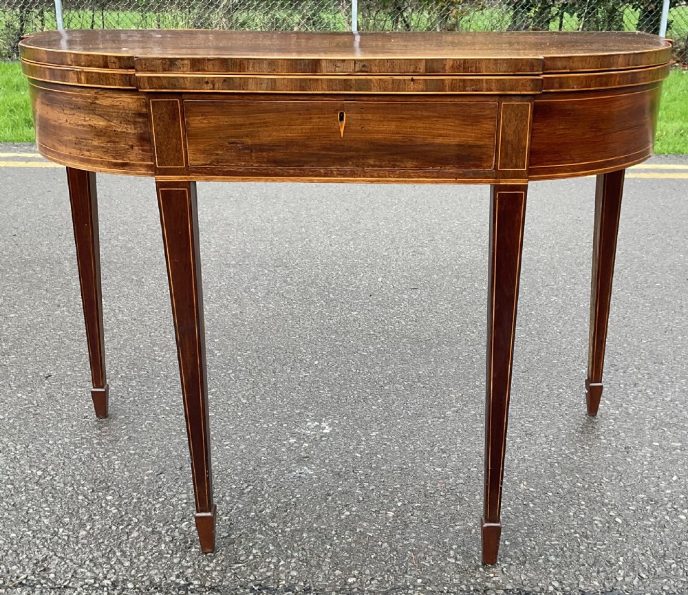 A Georgian demilune mahogany foldover tea table featuring boxwood stringing inlay, a central keyhole escutcheon, and four tapered square legs.