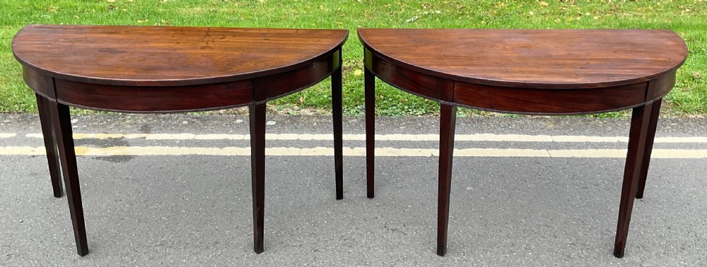A pair of Georgian dark mahogany demilune console tables with semi-circular tops and tapered square legs.