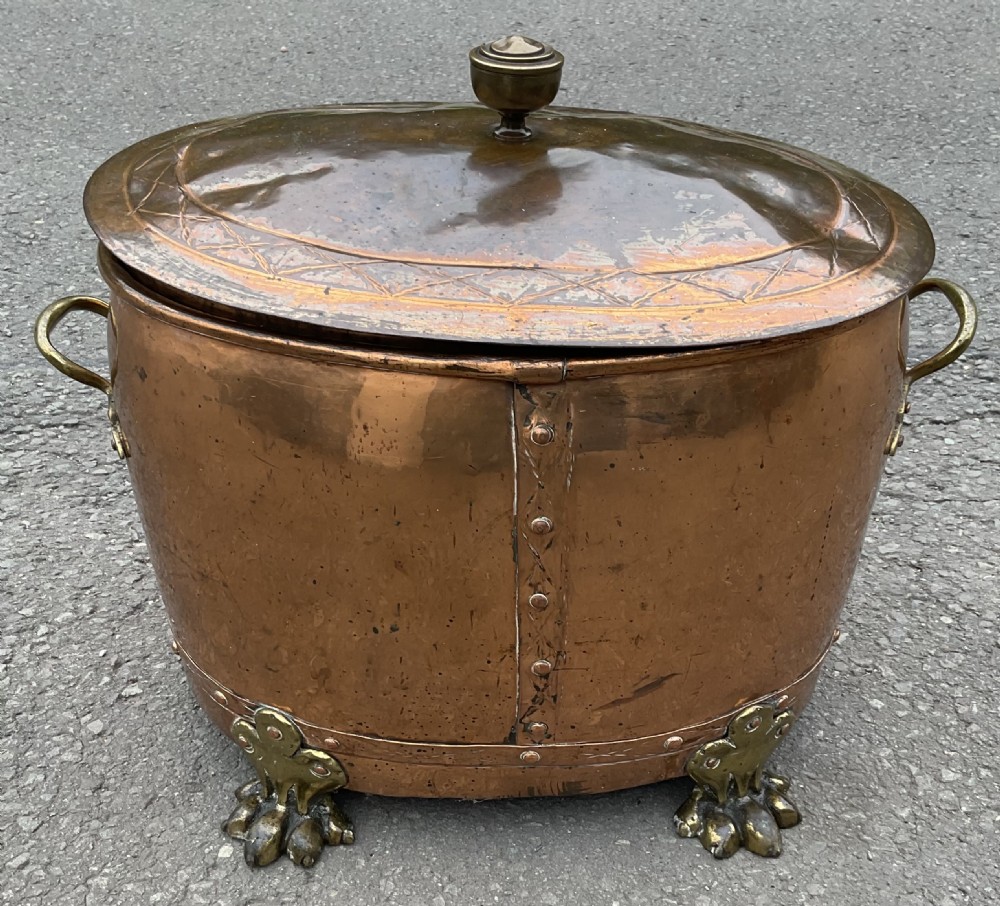 An antique oval copper log bin with brass claw feet, side handles, riveted seams, and an incised brass lid with a top finial.