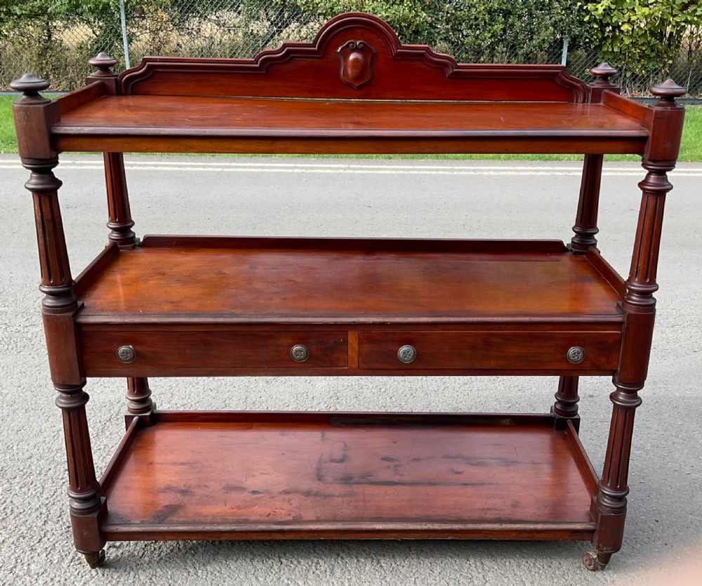 A Victorian three-tier mahogany dumb waiter with fluted turned supports, two frieze drawers, a carved crest backrail, and brass castors.