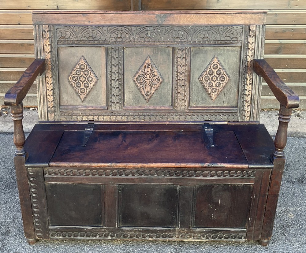 A dark carved oak hall settle featuring geometric lozenge panel carvings, turned arm supports, and a hinged lid storage seat.