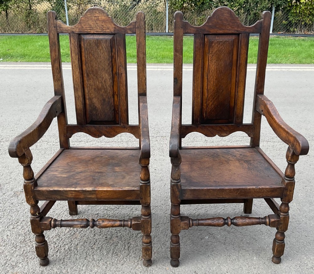 A pair of Tudor style dark oak armchairs featuring panelled backs, shaped open armrests, and turned front legs.