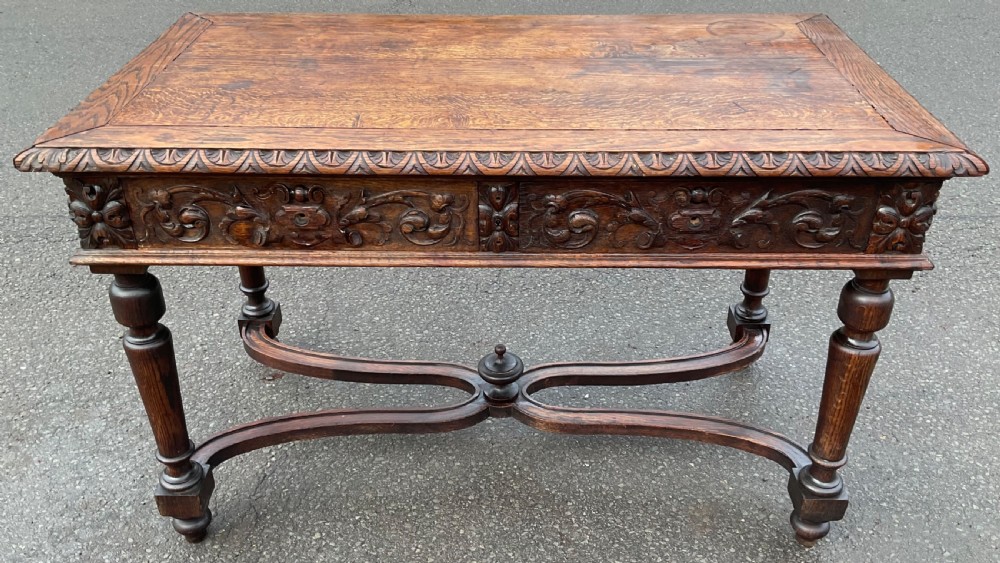 A Victorian dark oak writing table featuring deeply carved foliate friezes, two front drawers, turned legs, and a curved stretcher base with a central finial.