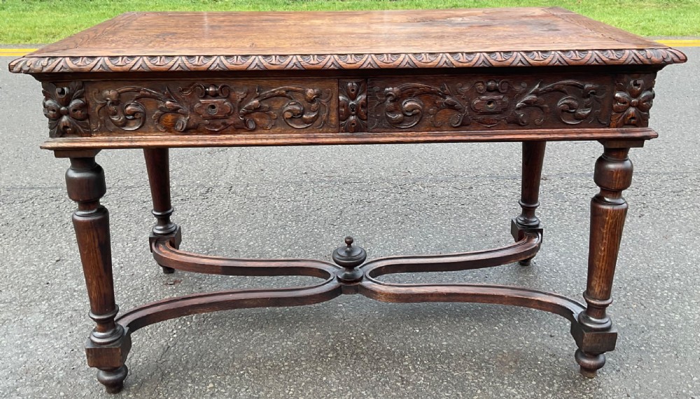 A Victorian dark oak writing table featuring deeply carved foliate friezes, two front drawers, turned legs, and a curved stretcher base with a central finial. View II