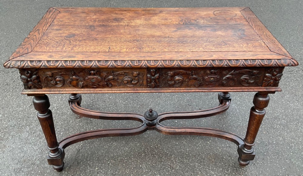 A Victorian dark oak writing table featuring deeply carved foliate friezes, two front drawers, turned legs, and a curved stretcher base with a central finial. View III