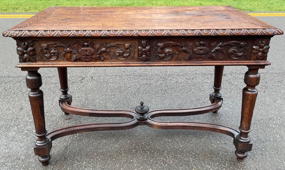 A Victorian dark oak writing table featuring deeply carved foliate friezes, two front drawers, turned legs, and a curved stretcher base with a central finial. View IV