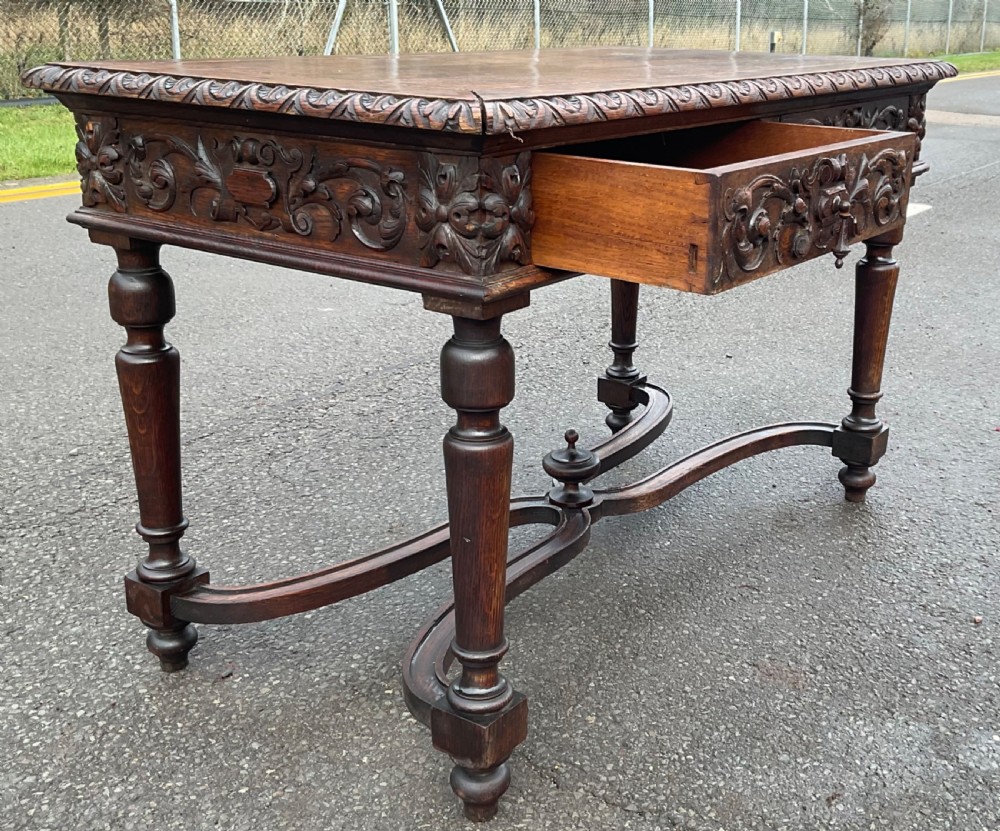 A Victorian dark oak writing table featuring deeply carved foliate friezes, two front drawers, turned legs, and a curved stretcher base with a central finial. View V