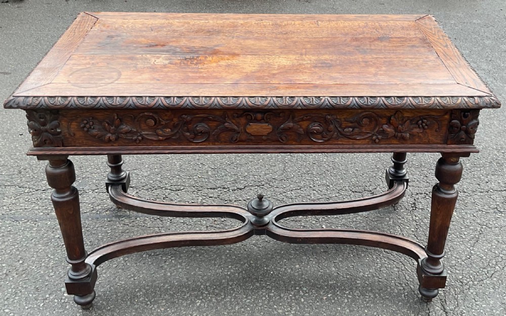 A Victorian dark oak writing table featuring deeply carved foliate friezes, two front drawers, turned legs, and a curved stretcher base with a central finial. View IX