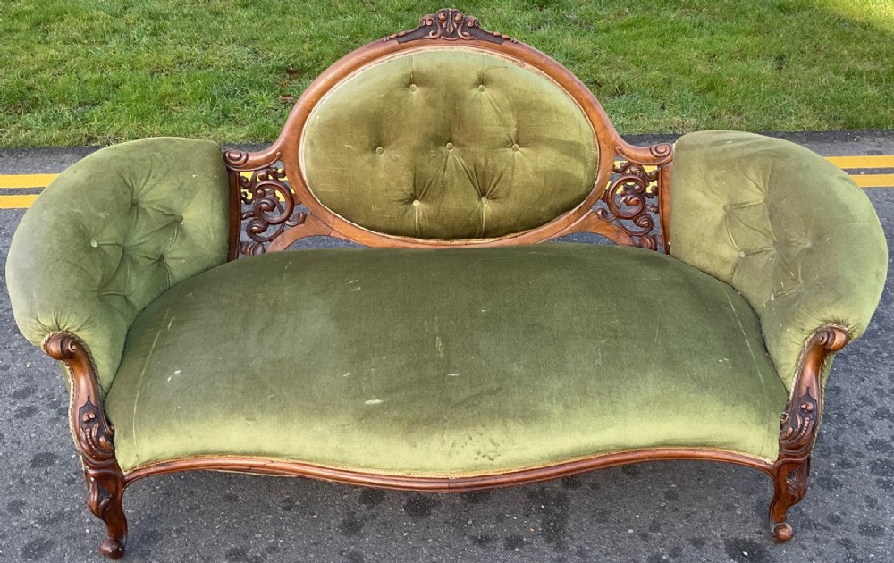 A Victorian walnut settee with an oval button-tufted backrest, carved wooden scrollwork, and olive green velvet upholstery.