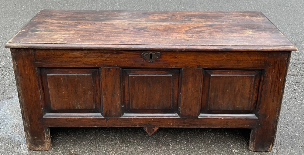 A dark oak antique blanket chest featuring three recessed front panels, a flat top hinged lid, and an iron escutcheon.