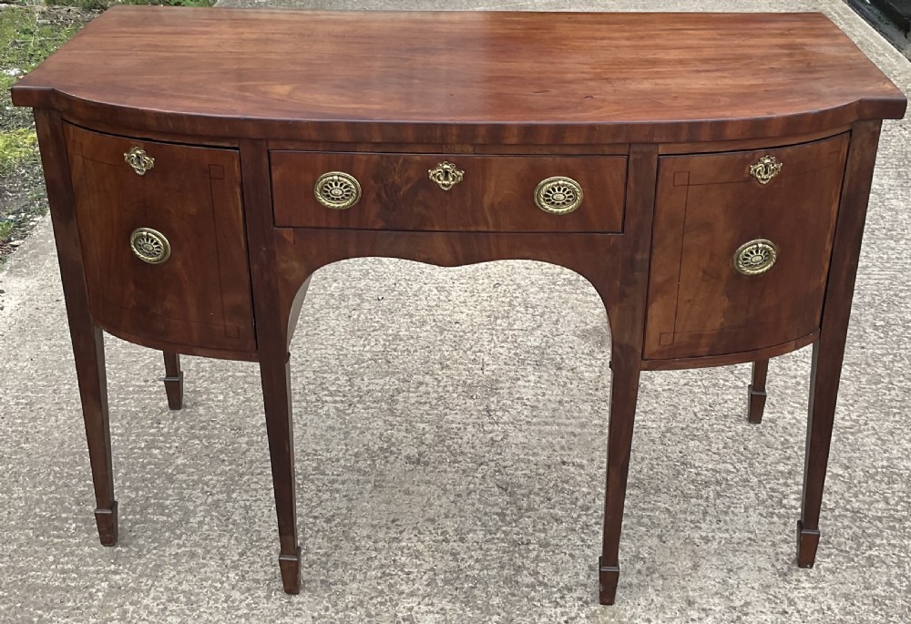 A Georgian mahogany bowfront sideboard featuring a central drawer flanked by two side cupboards, brass handles, and square tapered legs with spade feet. View IV
