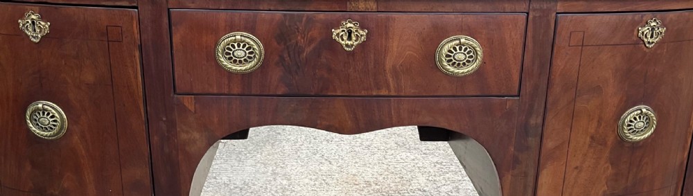 A Georgian mahogany bowfront sideboard featuring a central drawer flanked by two side cupboards, brass handles, and square tapered legs with spade feet. View VII