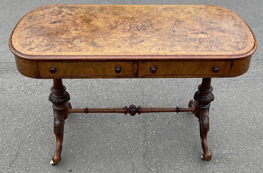 A Victorian burr walnut writing table featuring floral marquetry inlays, two frieze drawers, carved leg supports, a turned stretcher, and brass castors.