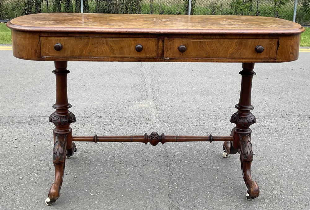A Victorian burr walnut writing table featuring floral marquetry inlays, two frieze drawers, carved leg supports, a turned stretcher, and brass castors. View III