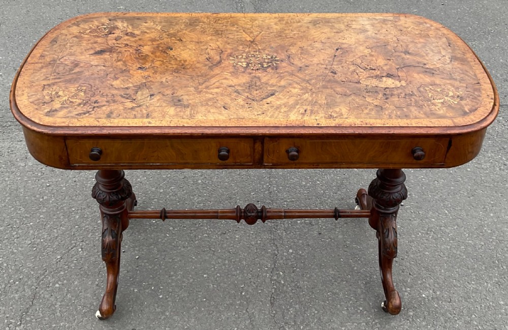 A Victorian burr walnut writing table featuring floral marquetry inlays, two frieze drawers, carved leg supports, a turned stretcher, and brass castors. View V