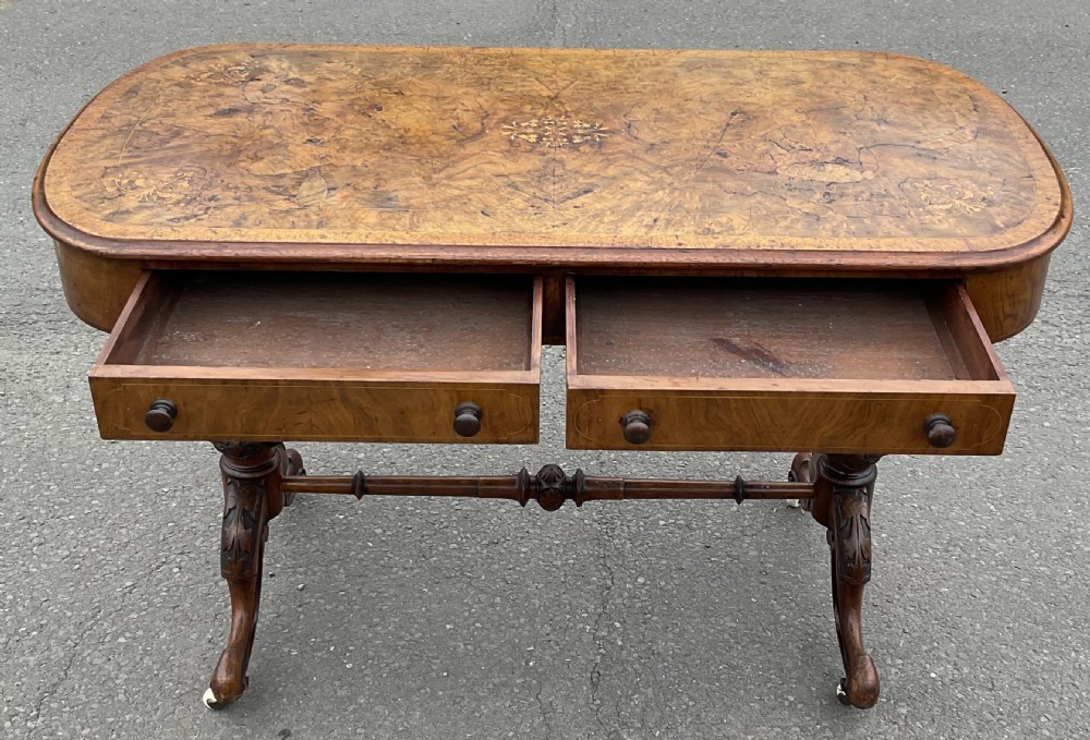 A Victorian burr walnut writing table featuring floral marquetry inlays, two frieze drawers, carved leg supports, a turned stretcher, and brass castors. View IX