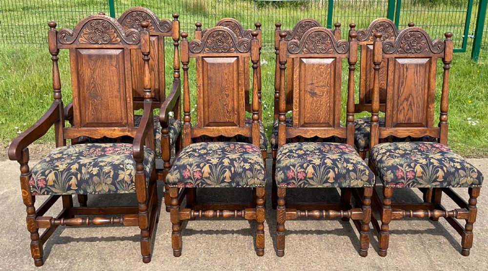 A set of eight Jacobean style oak dining chairs featuring carved arch crest rails, solid panel backs, turned stretchers, and floral tapestry upholstered seats.
