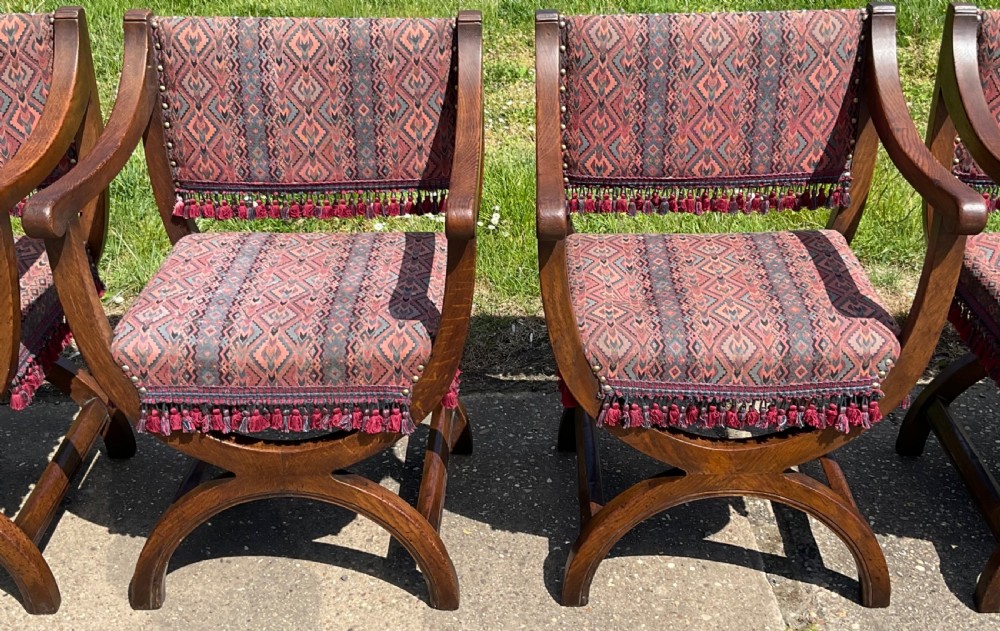 A set of six oak armchairs featuring cross-frame legs, curved armrests, red patterned geometric fabric, and fringed seat aprons. View V