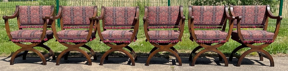 A set of six oak armchairs featuring cross-frame legs, curved armrests, red patterned geometric fabric, and fringed seat aprons. View VI
