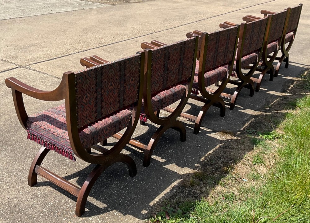 A set of six oak armchairs featuring cross-frame legs, curved armrests, red patterned geometric fabric, and fringed seat aprons. View VII