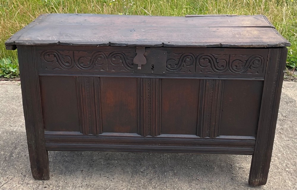 A dark carved oak blanket chest featuring carved top frieze scrolls, three recessed front panels, and extended stile legs.
