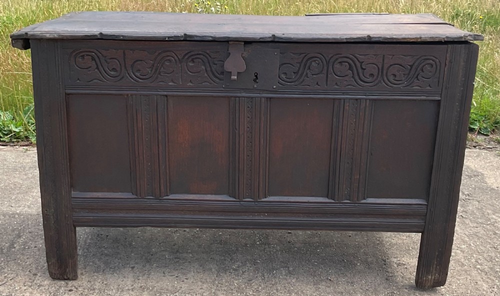 A dark carved oak blanket chest featuring carved top frieze scrolls, three recessed front panels, and extended stile legs. View II