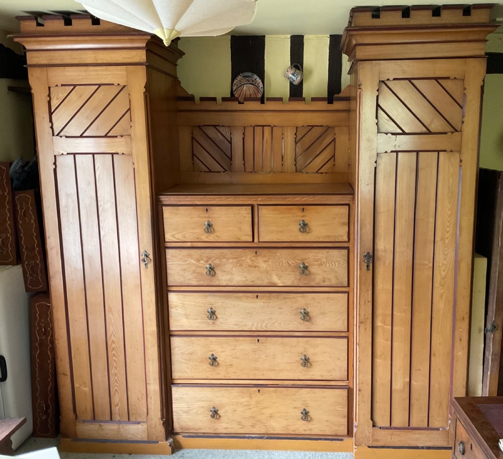 A large Aesthetic Movement pitch pine triple wardrobe featuring a central chest of drawers flanked by two tall cupboard doors with diagonal panel detailing and a castellated top cornice.