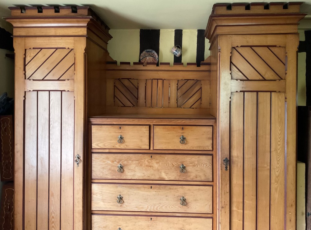 A large Aesthetic Movement pitch pine triple wardrobe featuring a central chest of drawers flanked by two tall cupboard doors with diagonal panel detailing and a castellated top cornice. View II