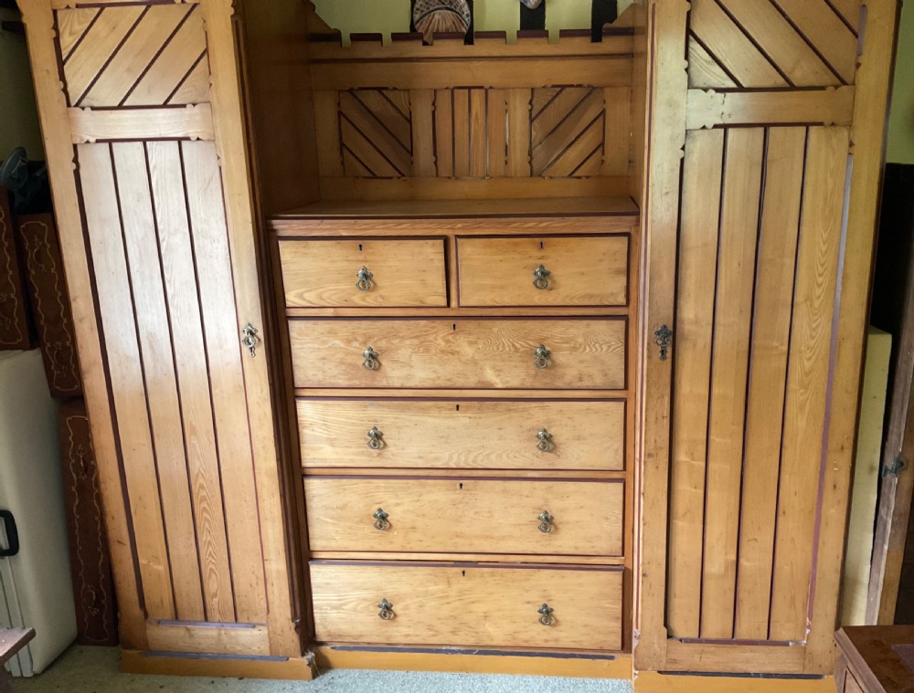 A large Aesthetic Movement pitch pine triple wardrobe featuring a central chest of drawers flanked by two tall cupboard doors with diagonal panel detailing and a castellated top cornice. View III