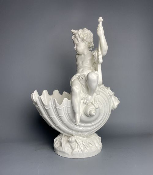 fig 1. A white glazed ceramic centrepiece featuring a cherub figure seated on a shell atop a naturalistic base.