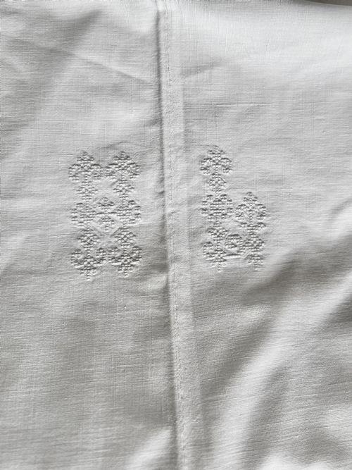 fig 1. Close-up view of a white handwoven homespun linen sheet featuring white hand-embroidered monograms along a central seam.