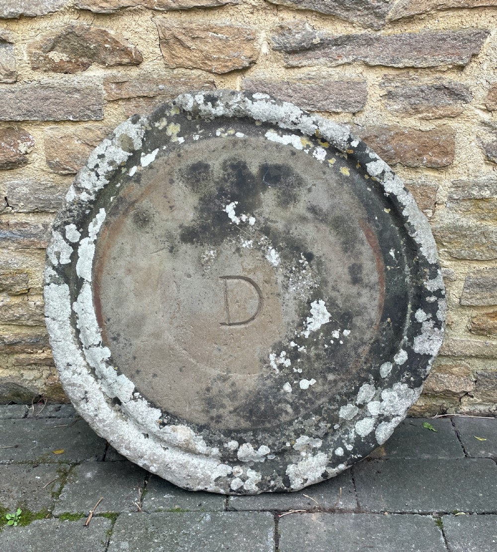 A circular 19th-century hand-carved grey limestone millstone base featuring a raised outer rim, lichen patina, and an incised letter D in the centre.