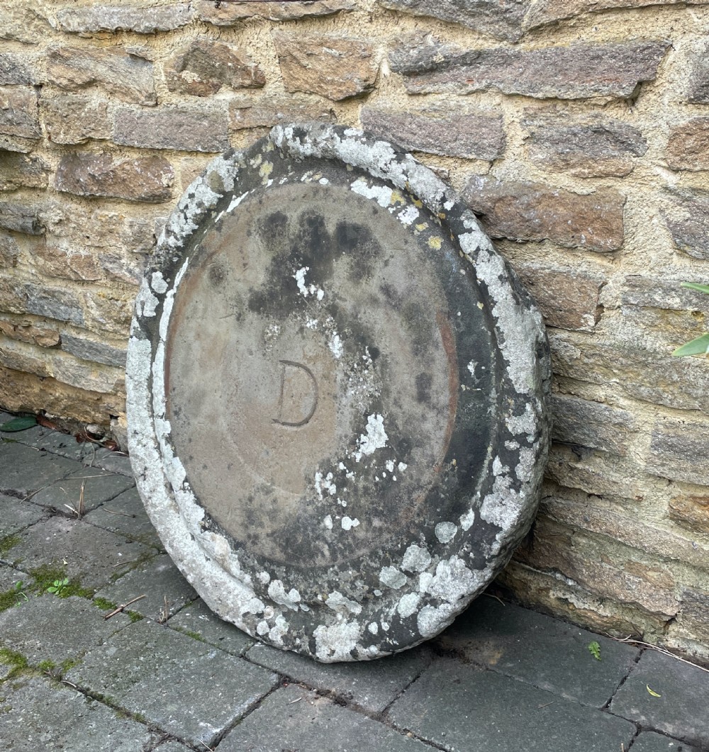 A circular 19th-century hand-carved grey limestone millstone base featuring a raised outer rim, lichen patina, and an incised letter D in the centre. View III