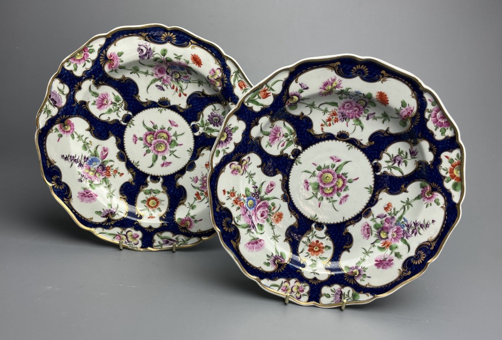 A pair of 18th-century circular porcelain plates featuring dark cobalt blue scale ground pattern with gilded rims, scalloped edges, and shaped white cartouches hand-painted with polychrome floral bouquets.