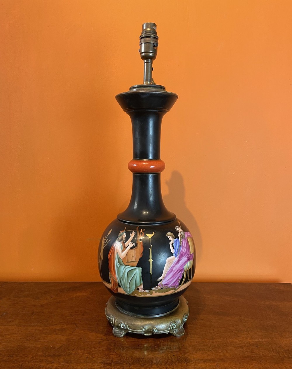 A tall black porcelain table lamp decorated with hand-painted classical Greek figures, a terracotta ring on the neck, and a cast brass base.
