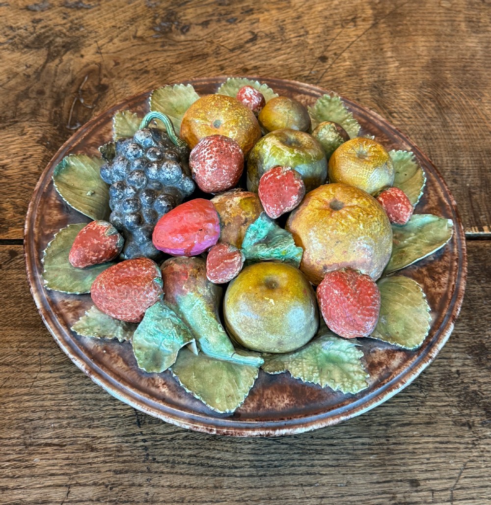 A circular 19th-century Portuguese majolica earthenware dish featuring high-relief ceramic fruit including grapes, strawberries, and oranges surrounded by green leaves. View V