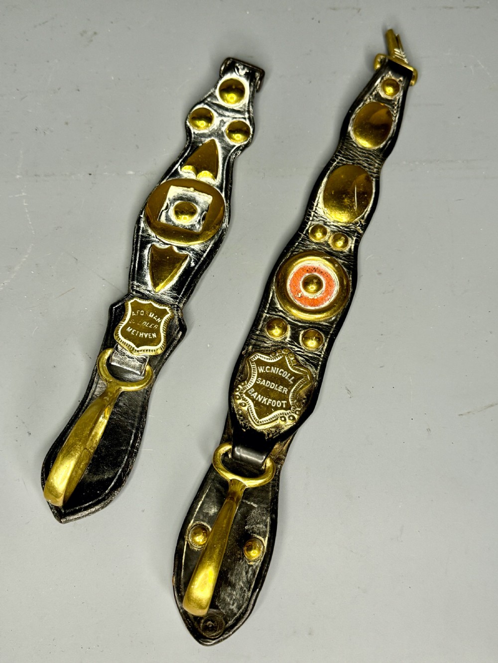 Two vertical Victorian leather martingales featuring polished cast brass amulets, stamped maker shields, and brass hooks against a neutral background.
