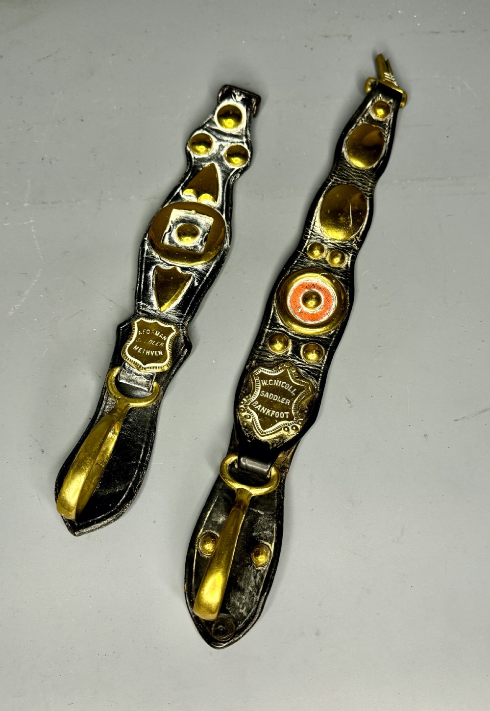 Two vertical Victorian leather martingales featuring polished cast brass amulets, stamped maker shields, and brass hooks against a neutral background. View II