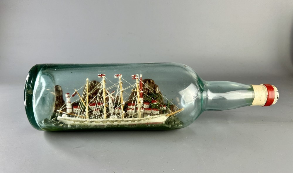 A vintage clear green glass bottle containing a detailed miniature four-masted sailing ship set against a coastal landscape with a lighthouse and windmill.