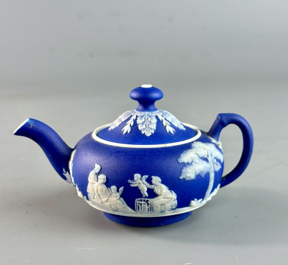 A miniature blue stoneware teapot featuring white classical relief figures and a leaf motif on the lid.