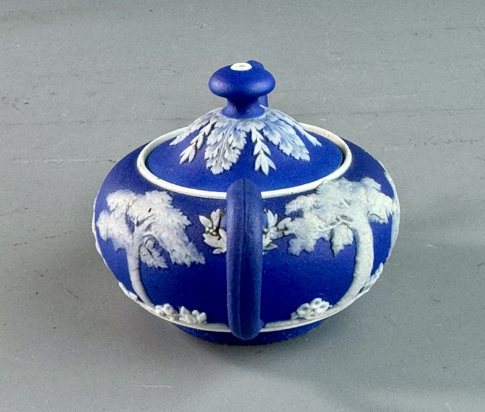 A miniature blue stoneware teapot featuring white classical relief figures and a leaf motif on the lid. View II