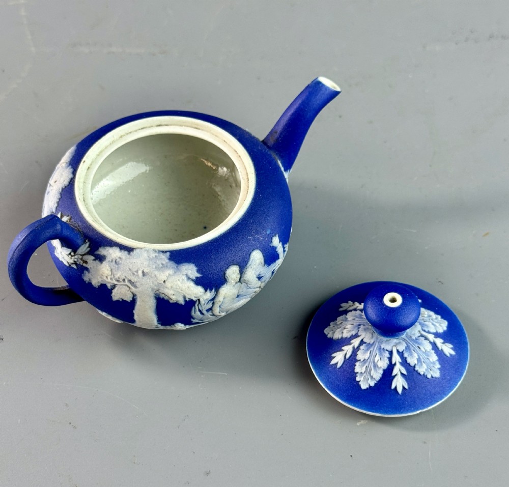 A miniature blue stoneware teapot featuring white classical relief figures and a leaf motif on the lid. View IV