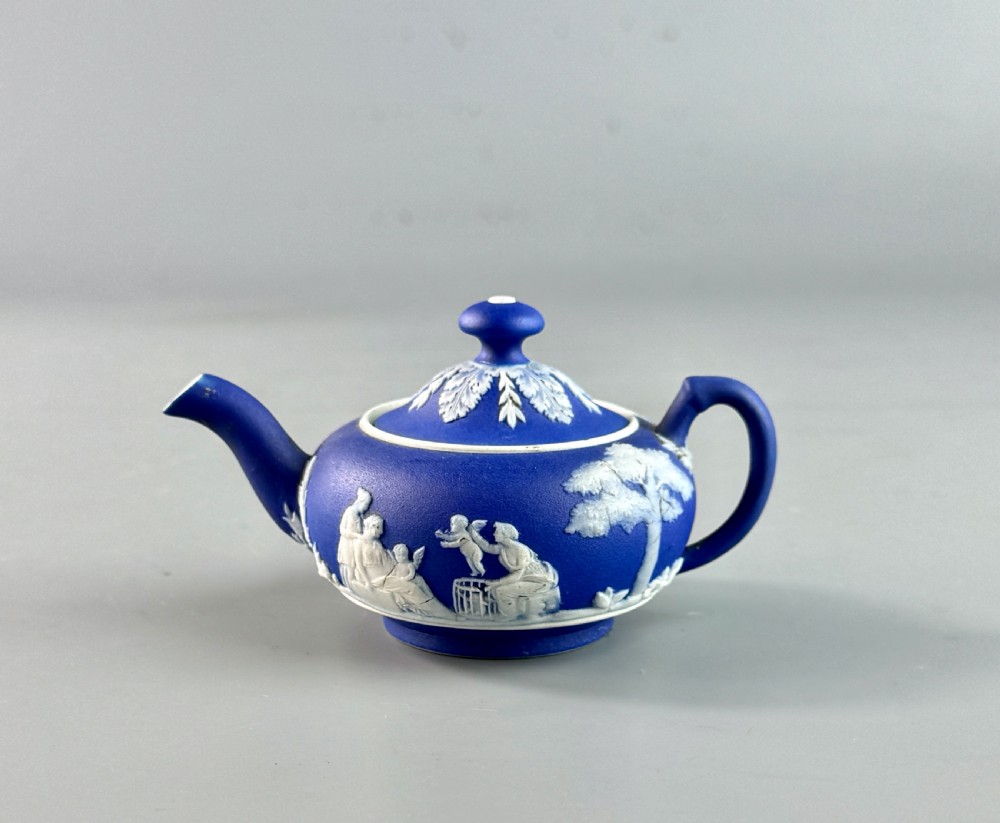A miniature blue stoneware teapot featuring white classical relief figures and a leaf motif on the lid. View V
