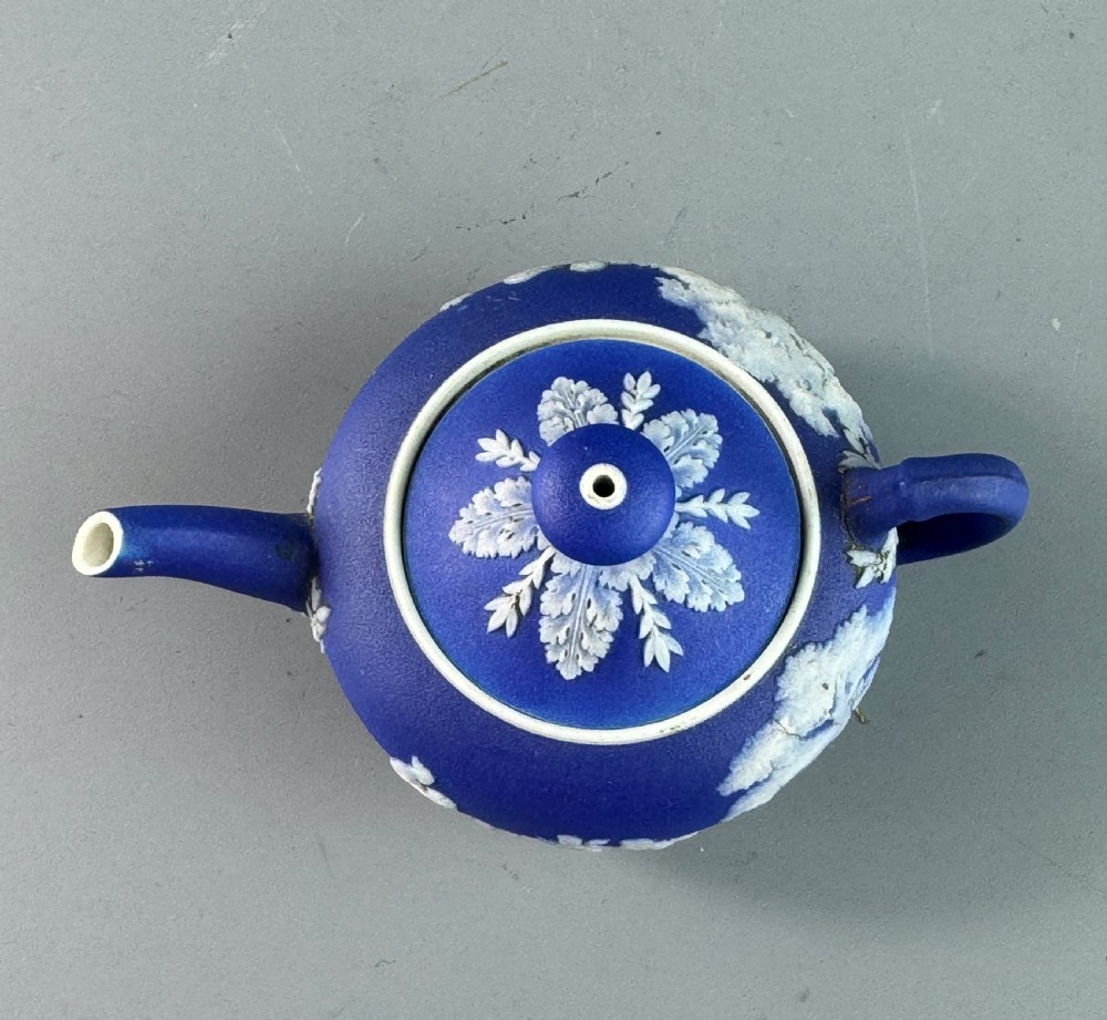 A miniature blue stoneware teapot featuring white classical relief figures and a leaf motif on the lid. View VII