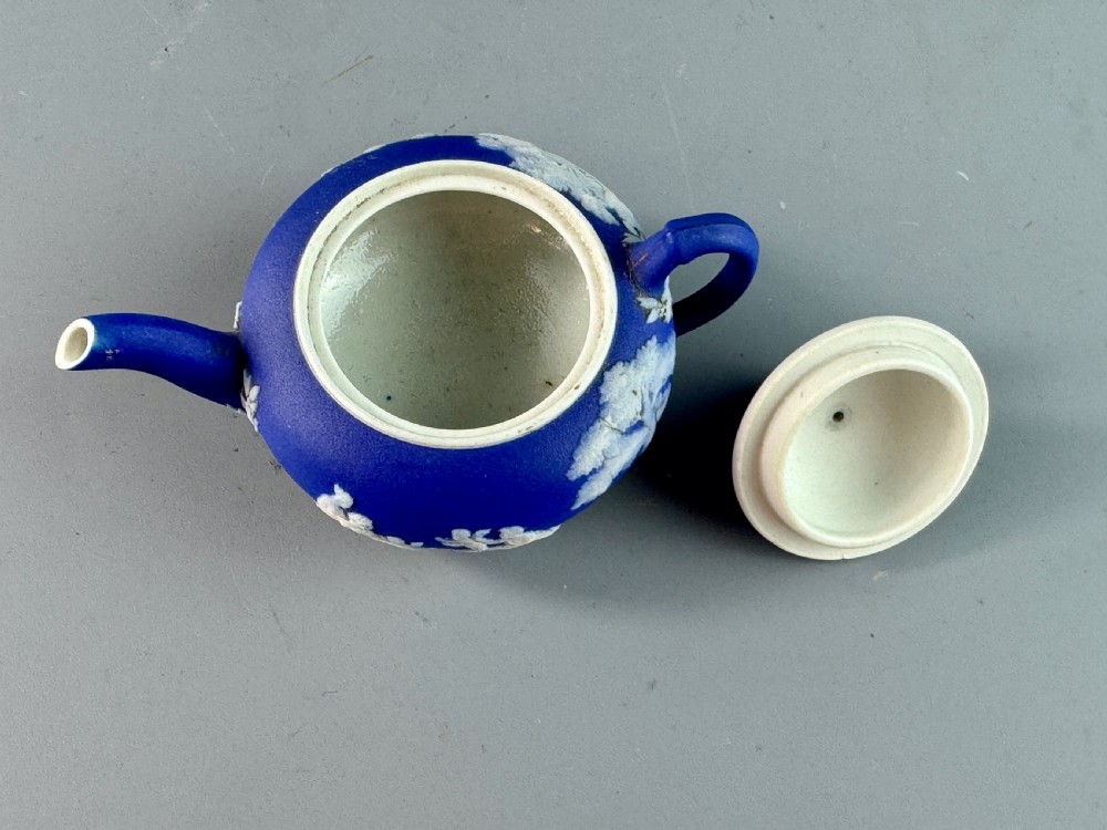 A miniature blue stoneware teapot featuring white classical relief figures and a leaf motif on the lid. View VIII