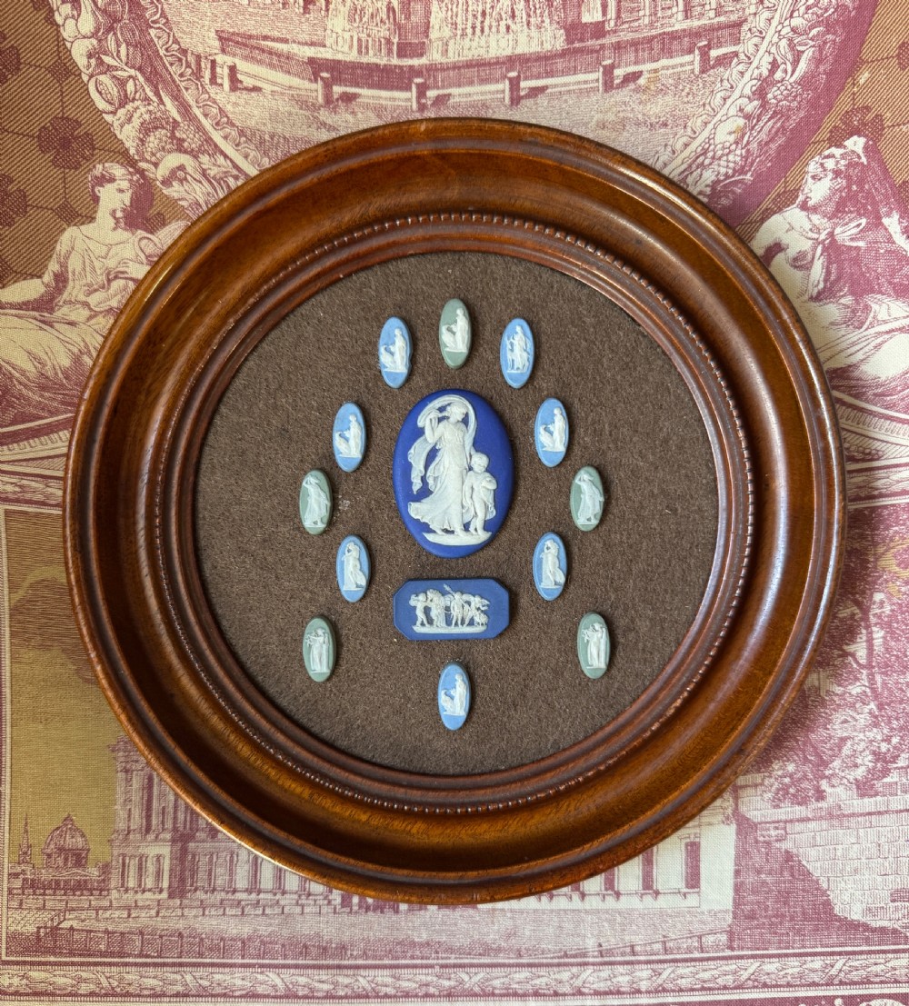 A circular wooden frame displaying fourteen miniature blue and sage green jasperware cameos with white classical figures mounted on brown fabric.