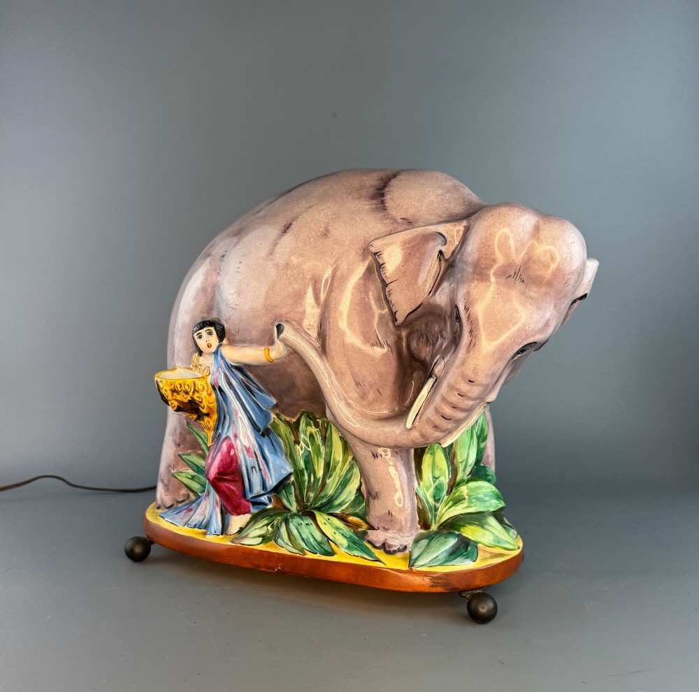 An Art Deco glazed porcelain perfume lamp depicting a mauve elephant alongside a female figure in blue drapery, set on green foliage.