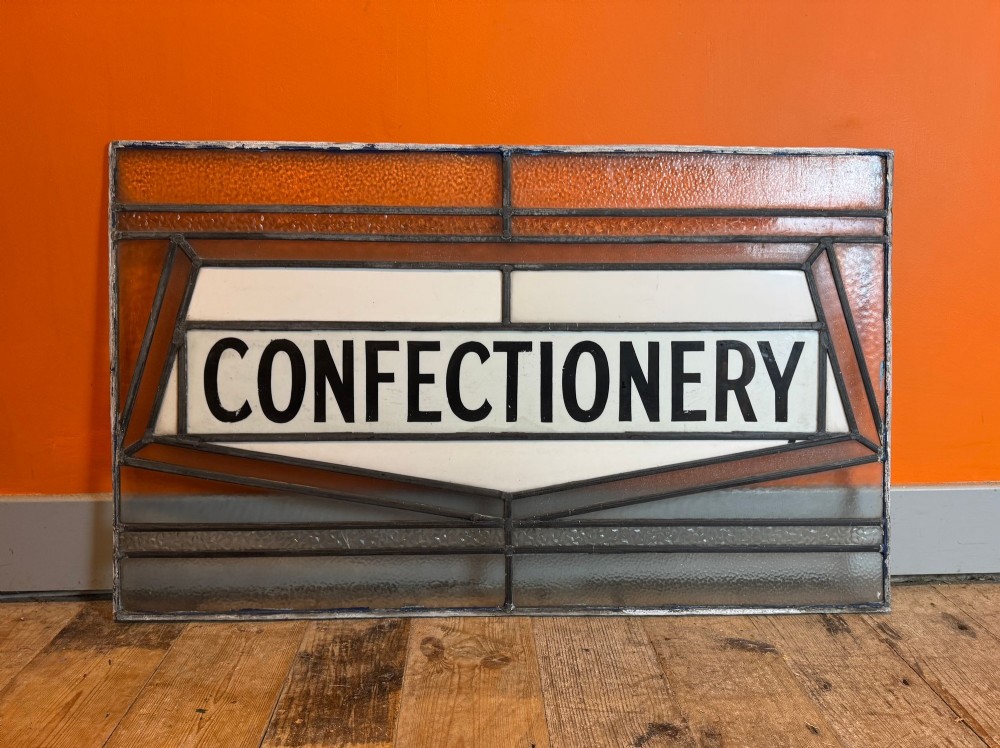 A rectangular leaded stained glass window panel with amber, clear, and white glass featuring the word CONFECTIONERY in bold black lettering.